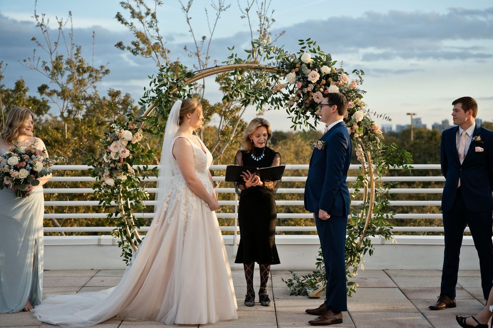 Bride and groom exchange vows under floral arch on rooftop venue at dusk with officiant and wedding party.