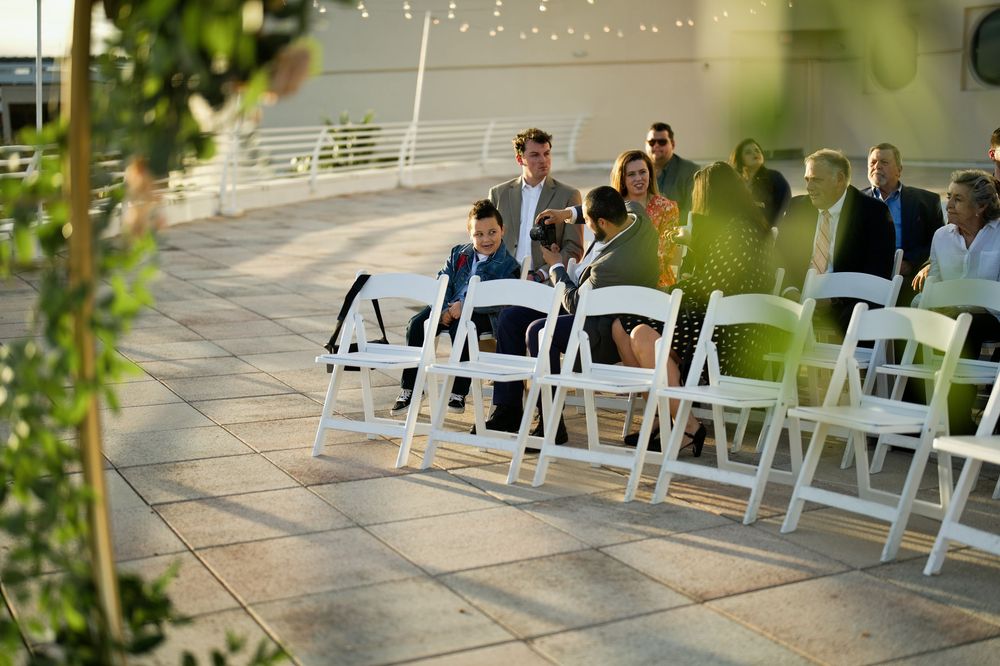 Guests seated in white folding chairs at an outdoor wedding ceremony on a patio with string lights overhead.