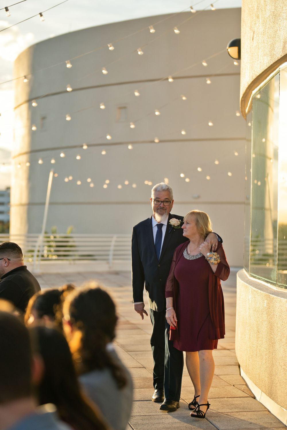 Elegant couple walking together at an outdoor wedding ceremony, man in navy suit with boutonniere, woman in burgundy dress.