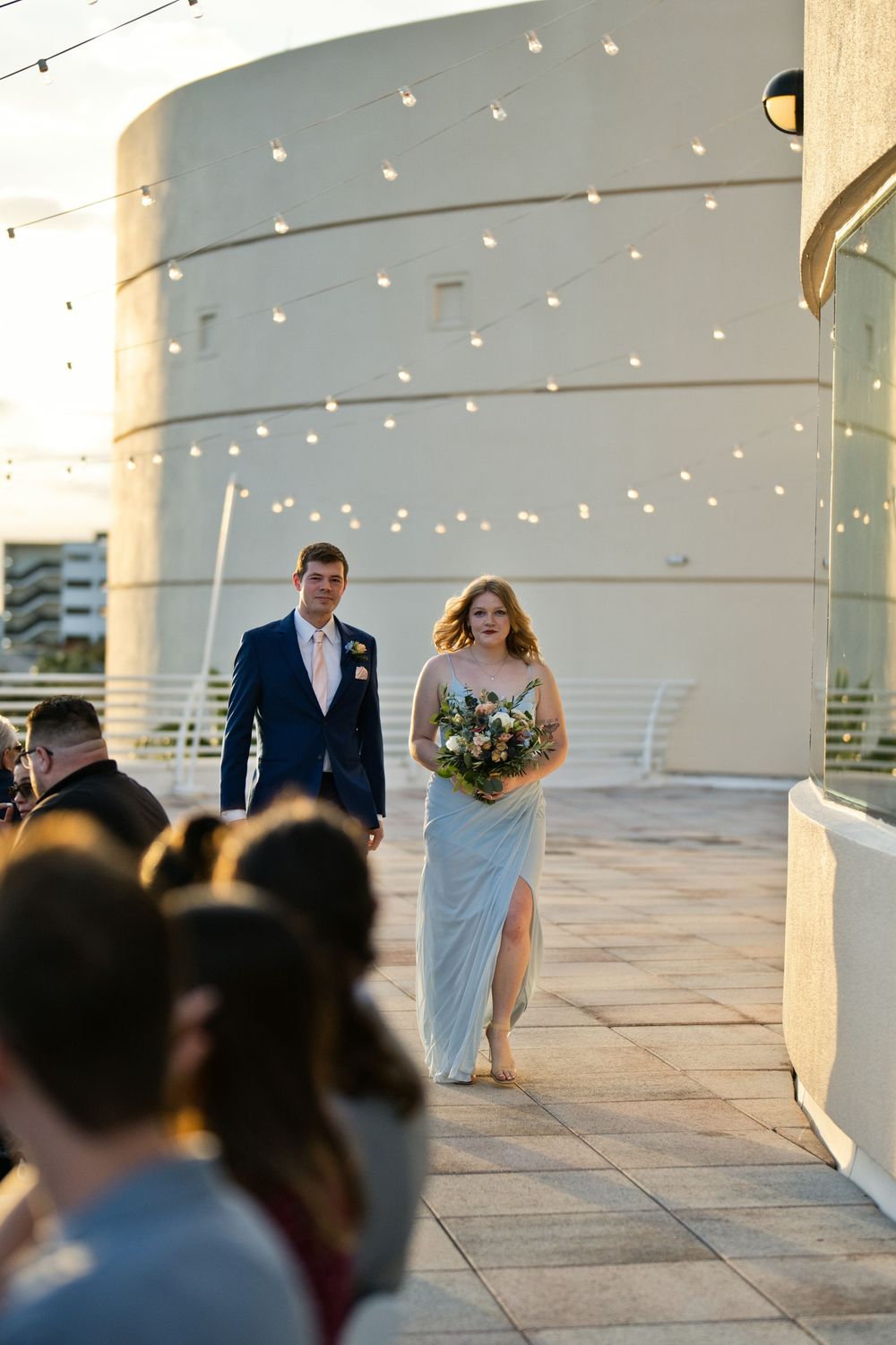 Bride in light blue gown holding bouquet walks with groomsman in navy suit along outdoor wedding venue.