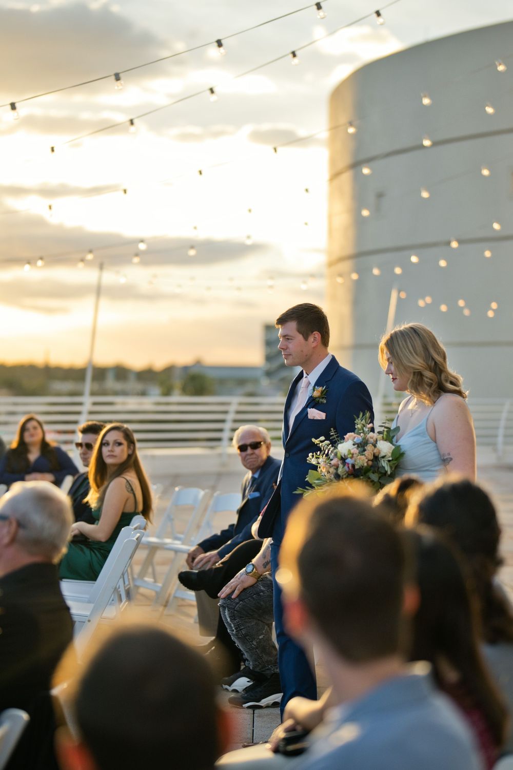 Groomsman escorts bridesmaid down outdoor rooftop wedding aisle at sunset with string lights and guests seated nearby.