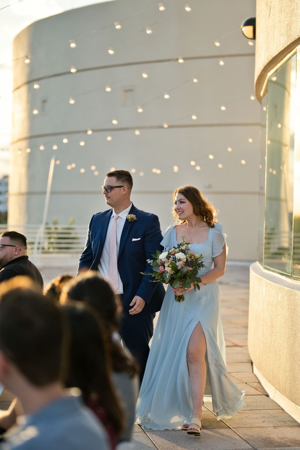 Bride in light blue gown holding bouquet walks with groom in navy suit during outdoor wedding ceremony at sunset.