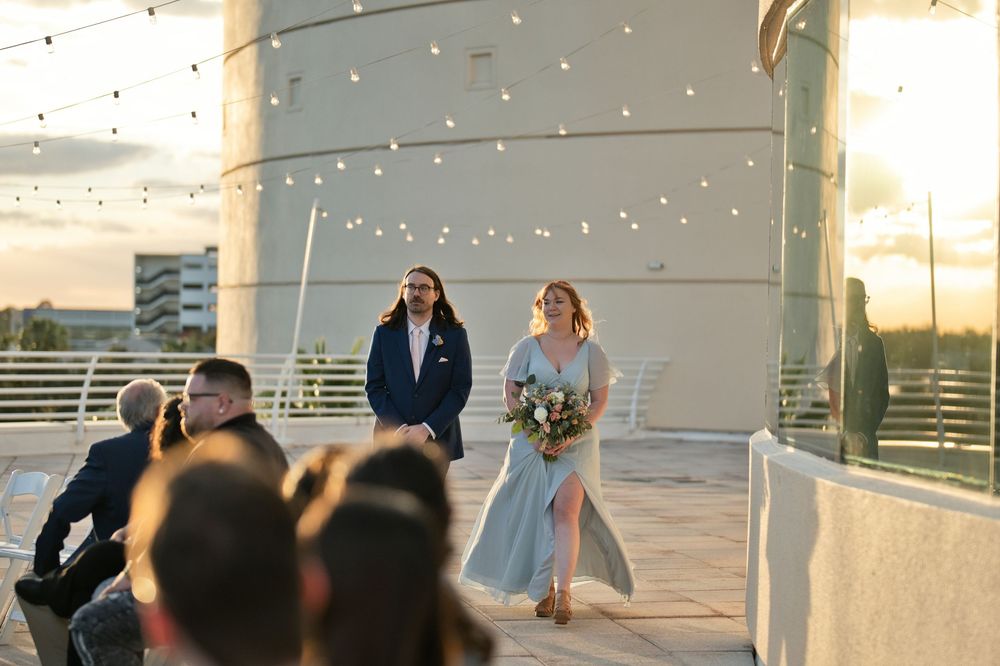 Bride in blue gown walks with escort on rooftop wedding venue at sunset with string lights.