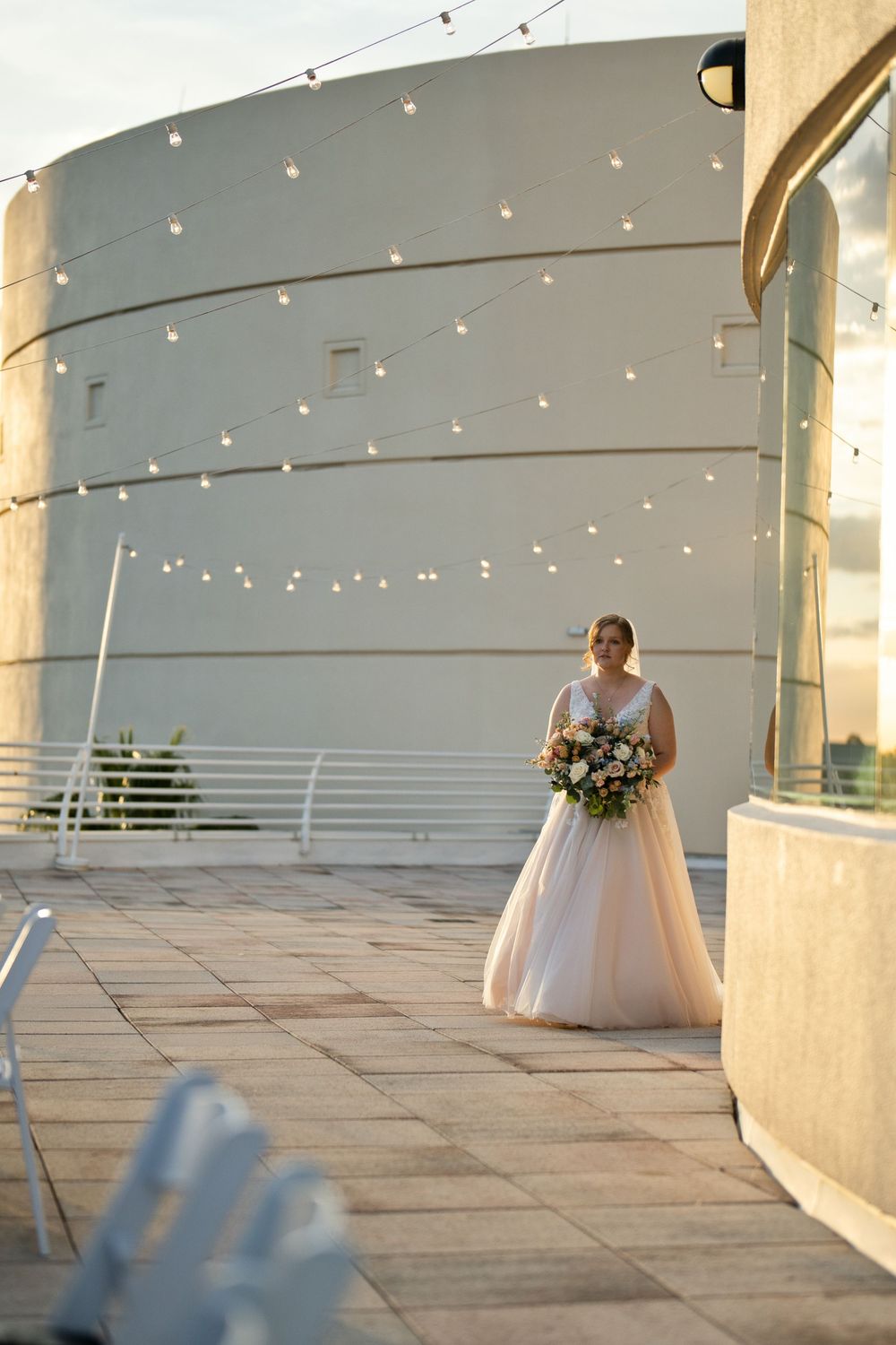 Bride in white gown holding floral bouquet stands on rooftop terrace with string lights and curved white building backdrop.