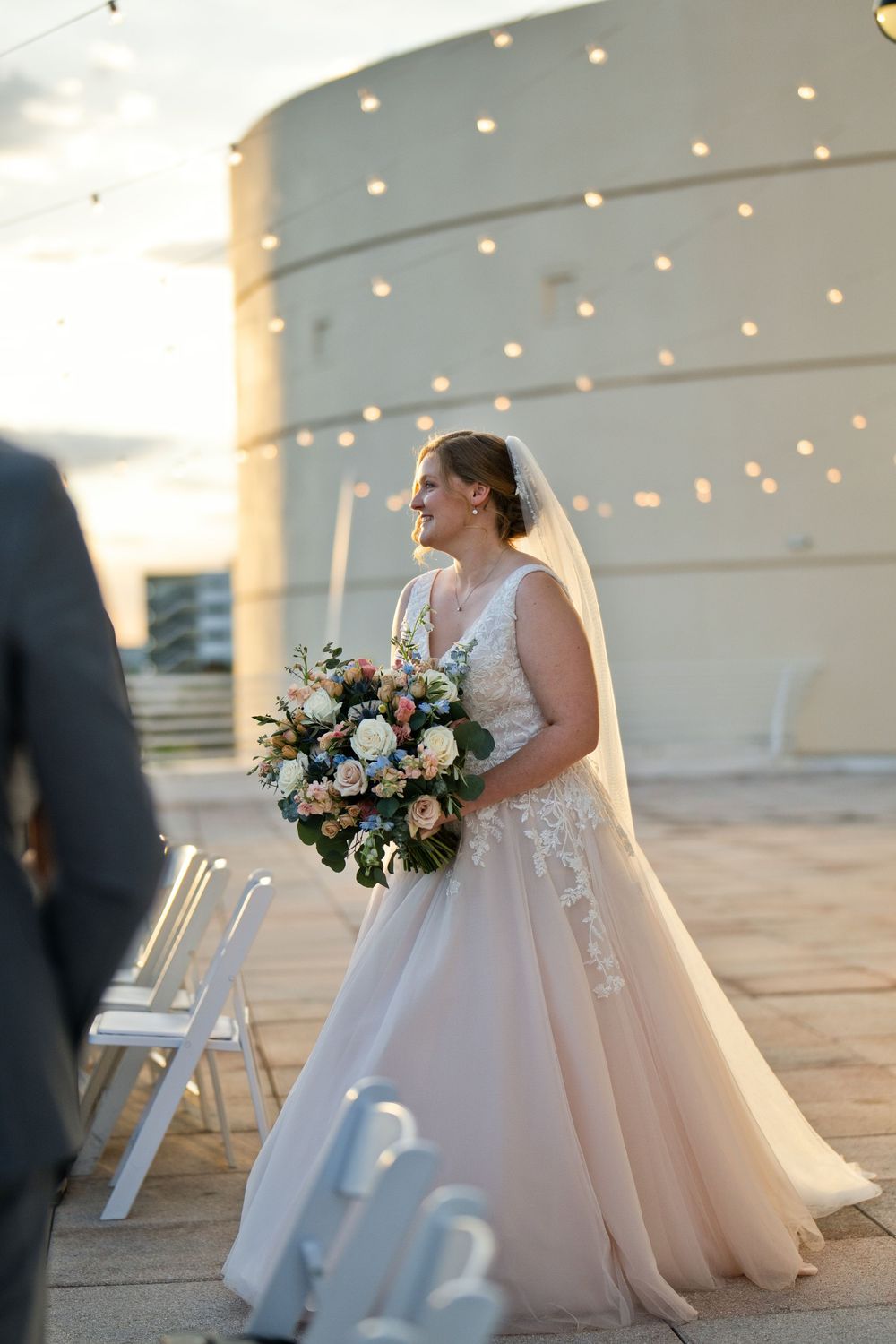 Smiling bride in blush lace ball gown holding floral bouquet walks toward altar at outdoor rooftop wedding ceremony.