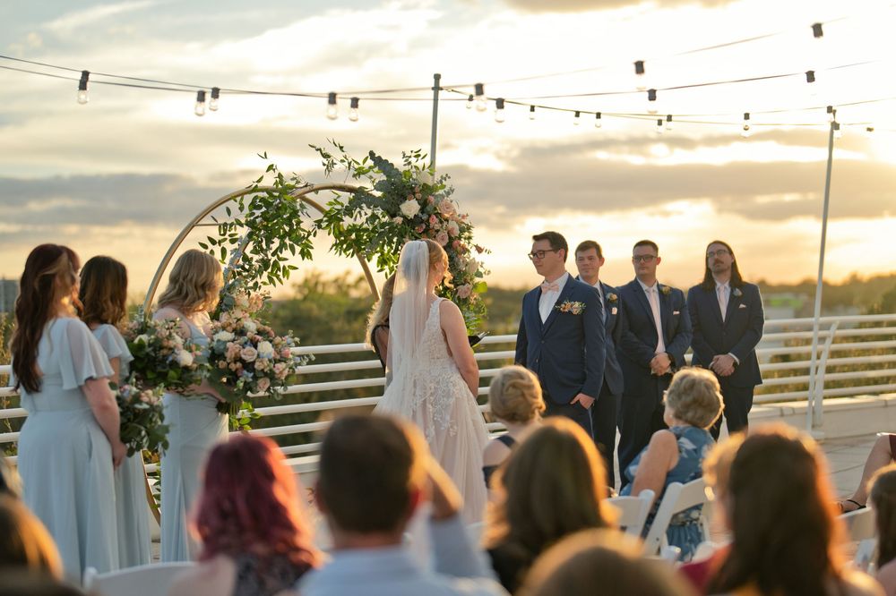 Outdoor rooftop wedding ceremony at sunset with couple at floral arch, string lights, and guests seated in rows.