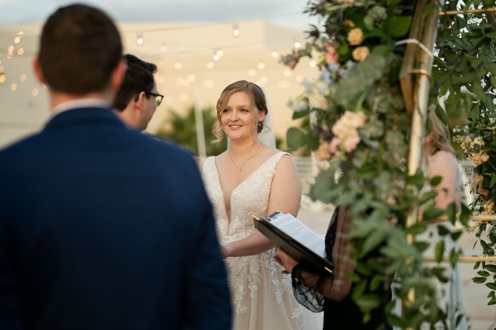 A bride smiles warmly at her groom during an outdoor wedding ceremony adorned with lush greenery and soft peach floral arrangements.