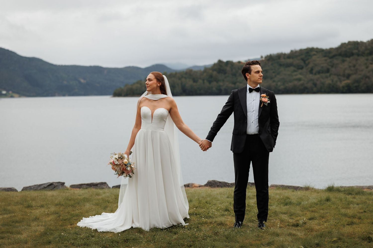Bride in white gown and groom in black tuxedo hold hands by a scenic lake with mountains in the background.