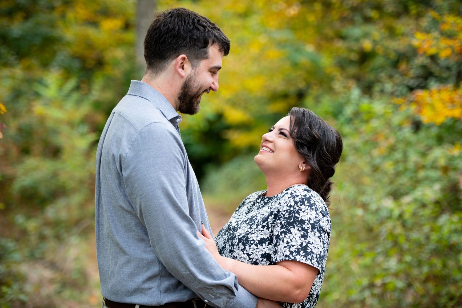 Couple sharing a romantic moment outdoors, smiling at each other surrounded by autumn foliage in a lush green setting.
