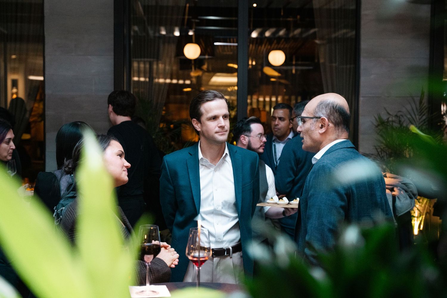 Professionals networking at an elegant indoor-outdoor cocktail party, holding wine glasses amid lush greenery.