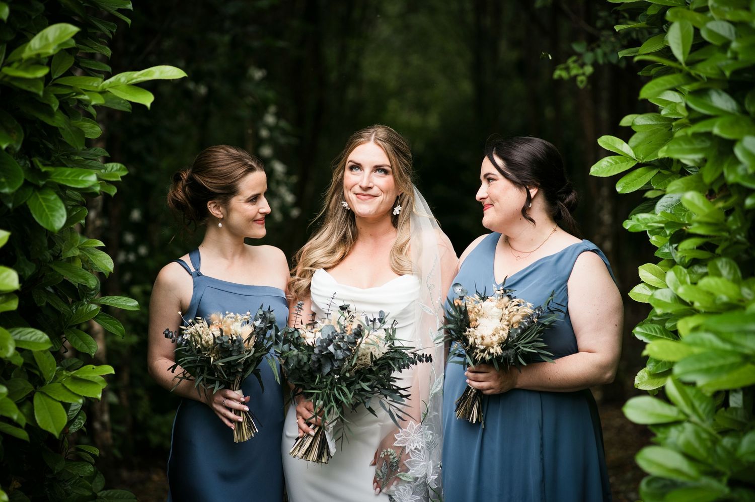 Bride with two bridesmaids in blue dresses holding bouquets of greenery and white flowers in a lush garden setting.