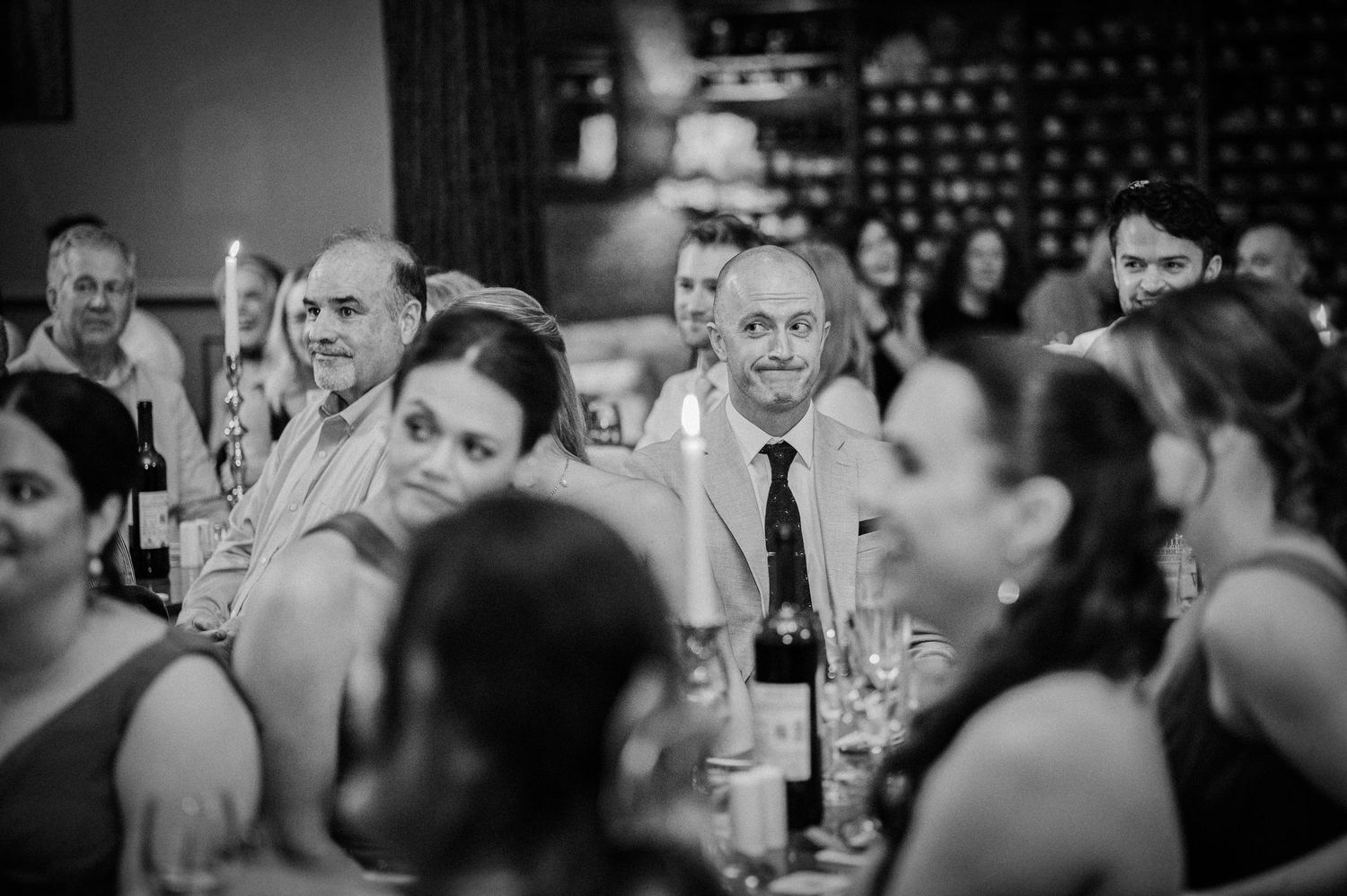 Wedding guests seated at dinner tables smile and watch speeches in a candid black and white reception photo.