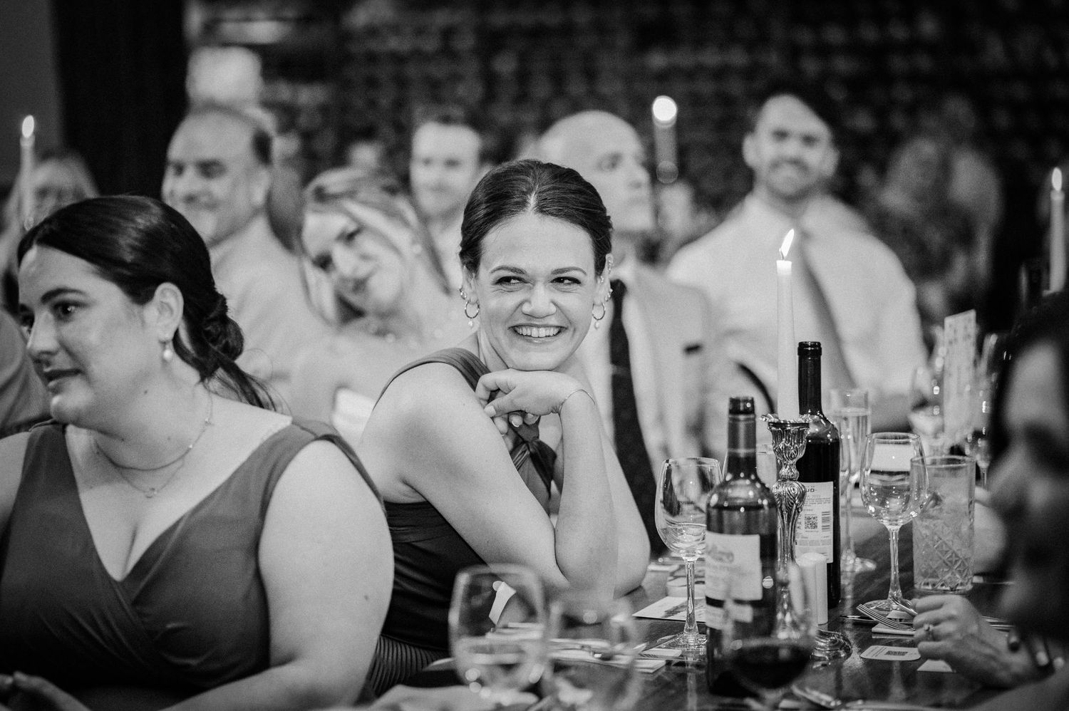 Black and white wedding reception photo of smiling guests seated at a decorated table with wine bottles and candles.
