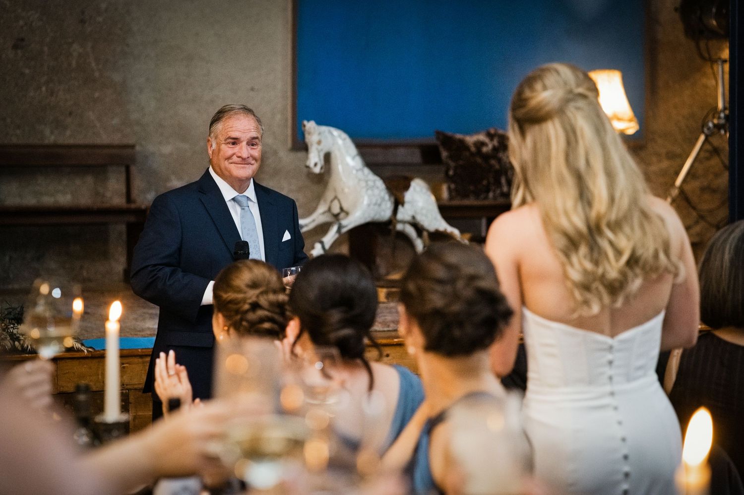 Man in navy suit smiling at wedding reception guests seated at candlelit table with white horse sculpture backdrop.