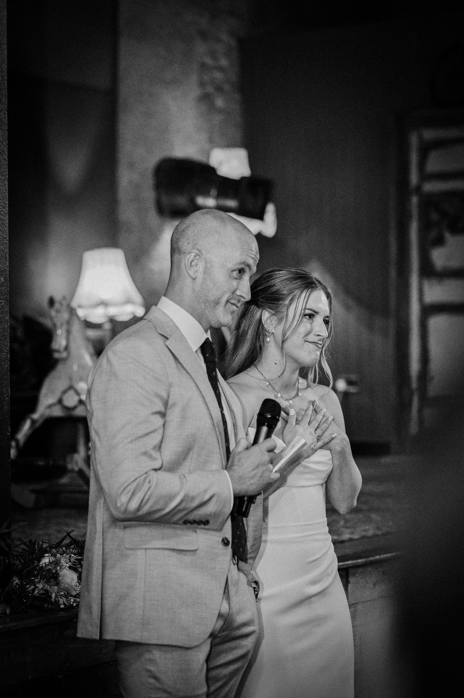 Emotional bride and groom listen to wedding speeches in a warmly lit reception venue.