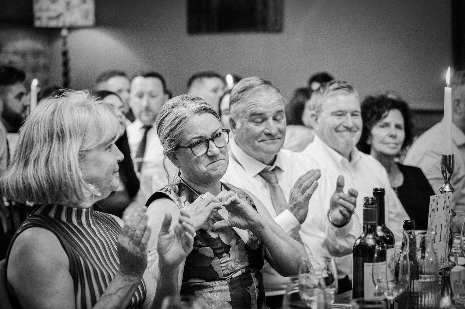Wedding guests clap and smile during speeches at an elegant candlelit reception dinner.