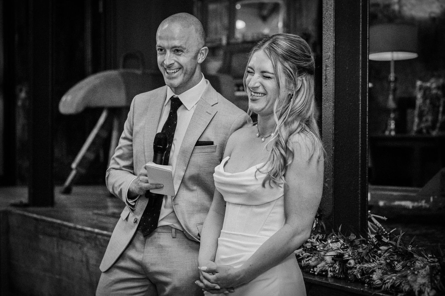 Bride and groom laugh together outside their wedding venue in black and white photo.