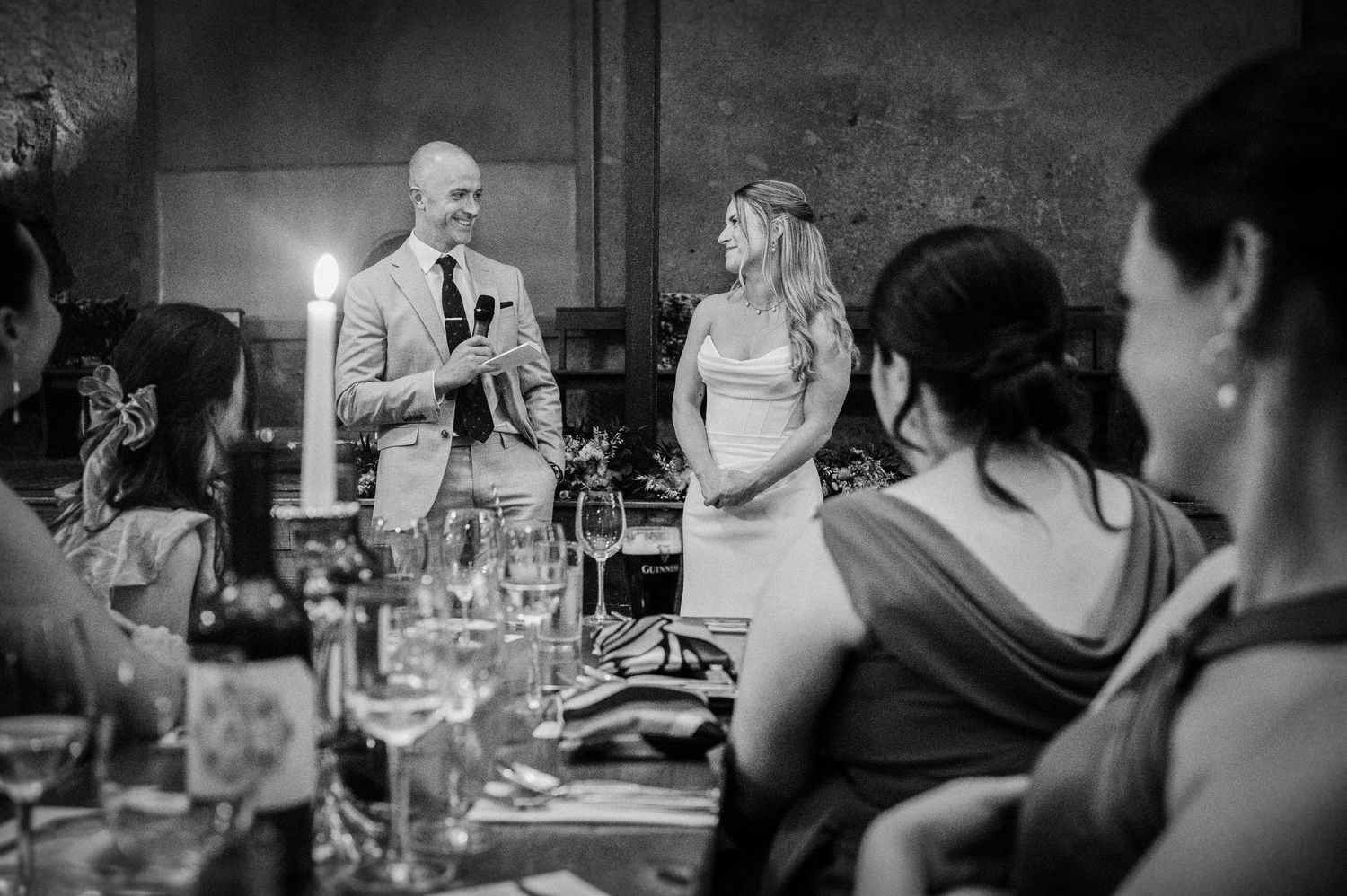 Groom gives speech beside smiling bride at candlelit wedding reception dinner table.