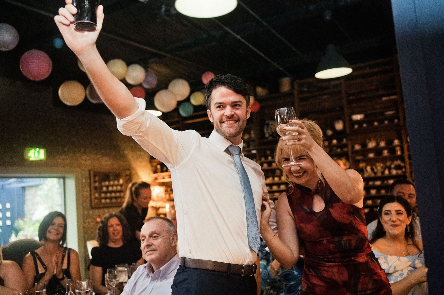 Man raising a glass in a toast with a woman at a lively wedding reception with colorful bokeh lights.