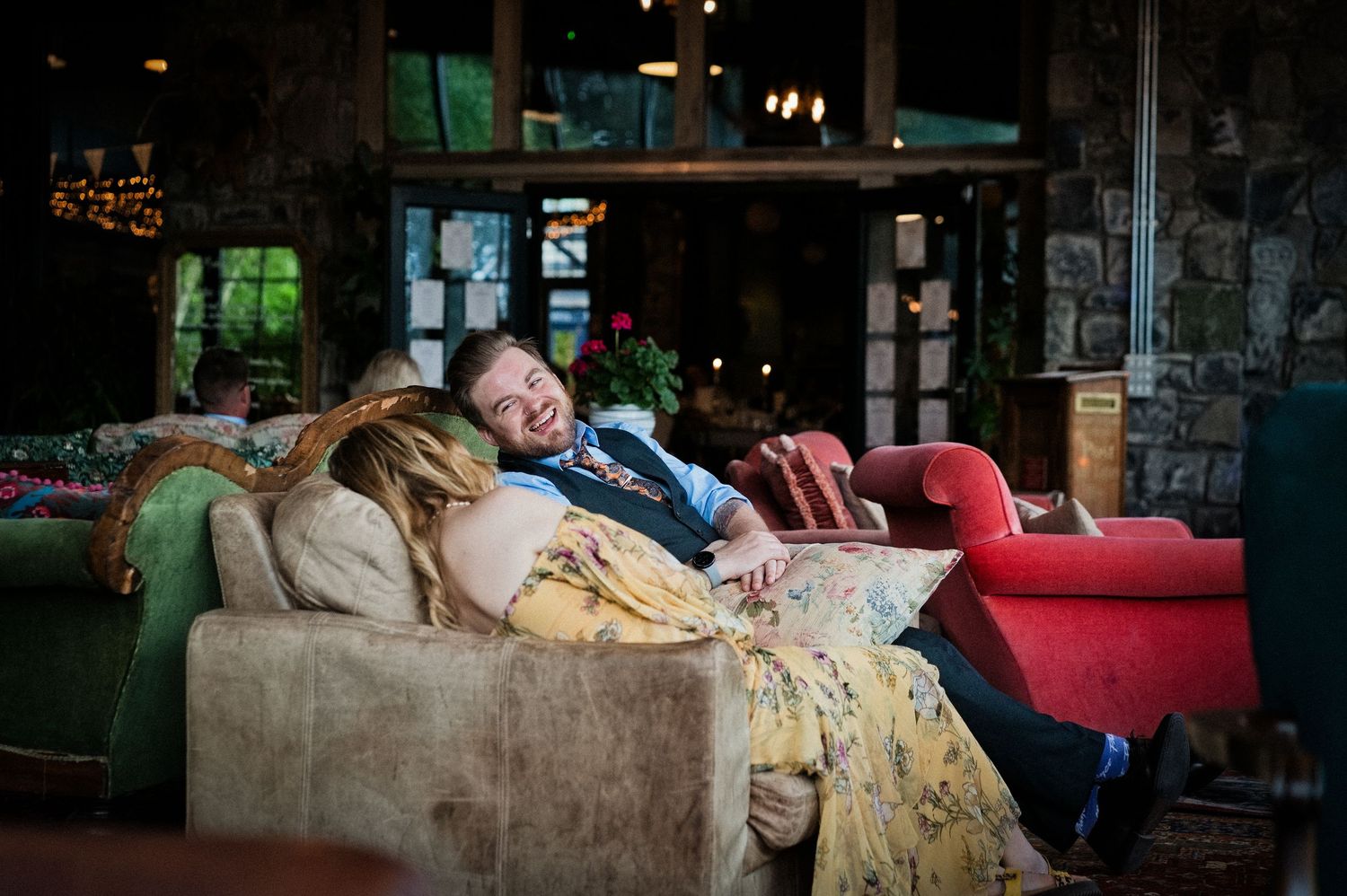 Happy couple relaxes together laughing on a vintage sofa in a rustic wedding venue.