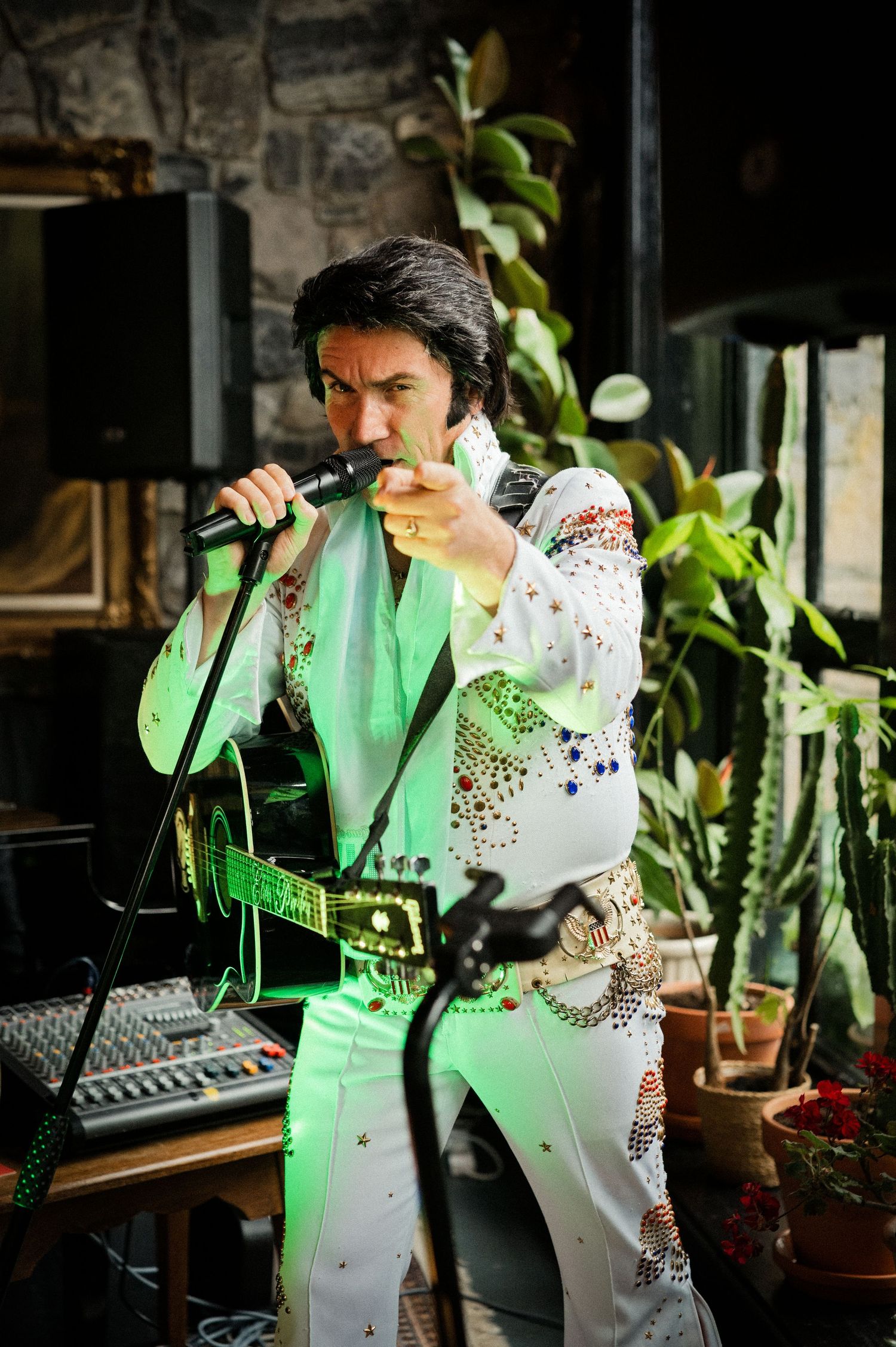 Elvis impersonator performs in a white jeweled jumpsuit, pointing at the crowd during a live wedding entertainment show.