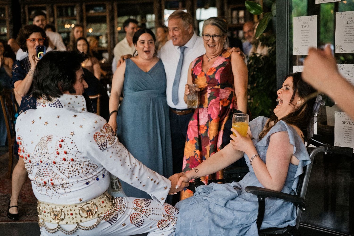 Wedding guests laughing with Elvis impersonator and woman in wheelchair at lively reception celebration.