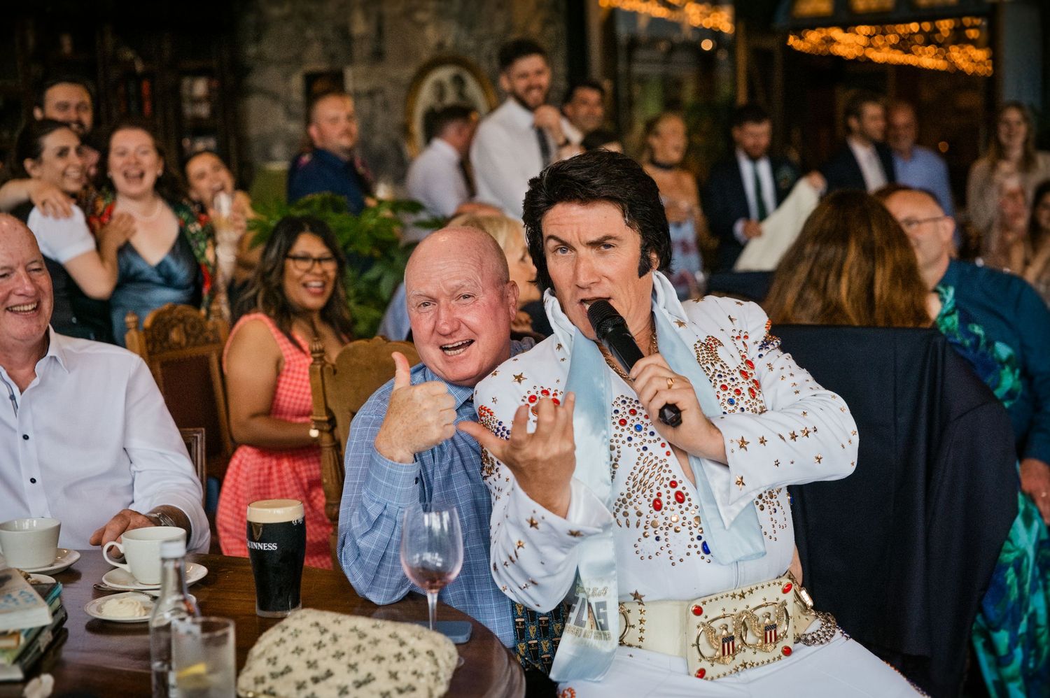 Elvis impersonator entertains wedding guests at their table, posing with a laughing guest during the reception celebration.