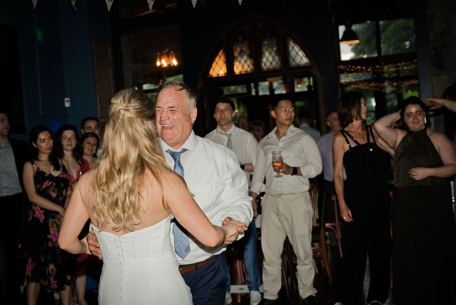 Bride and older man share a joyful dance at a wedding reception with guests watching in the background.