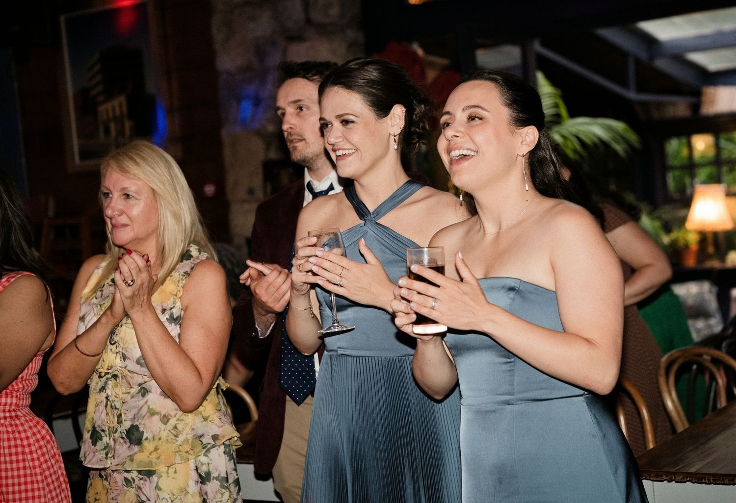 Women in blue bridesmaid dresses clapping and smiling at a wedding reception indoor venue.