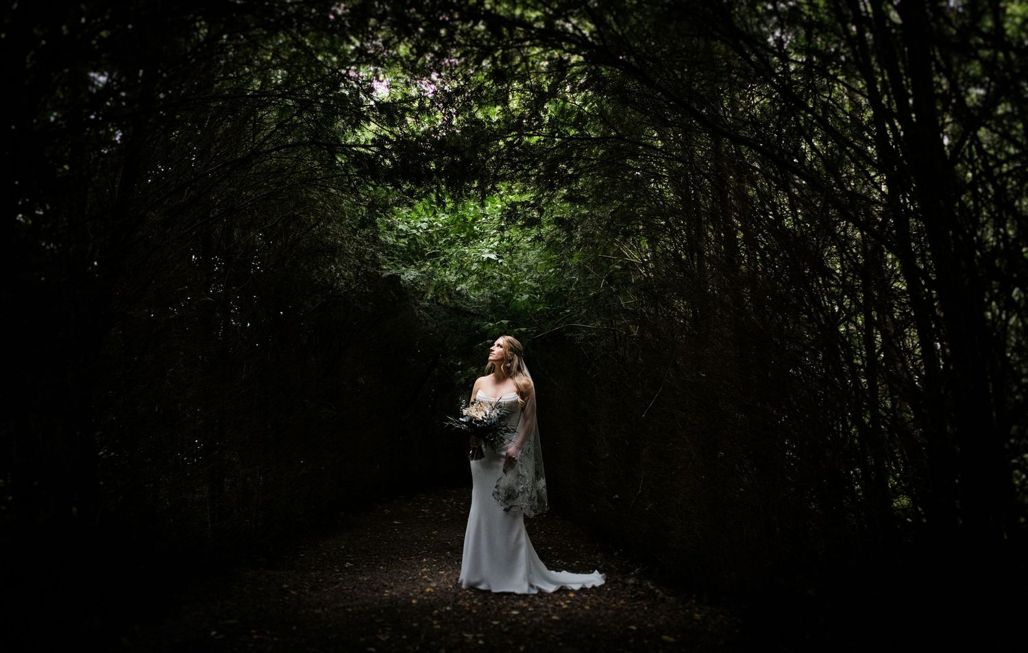 Bride in white gown standing in enchanting dark forest tunnel holding bouquet.