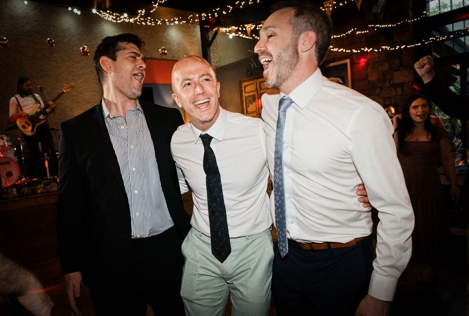 Three men in dress shirts and ties laugh together at a wedding reception venue with string lights.