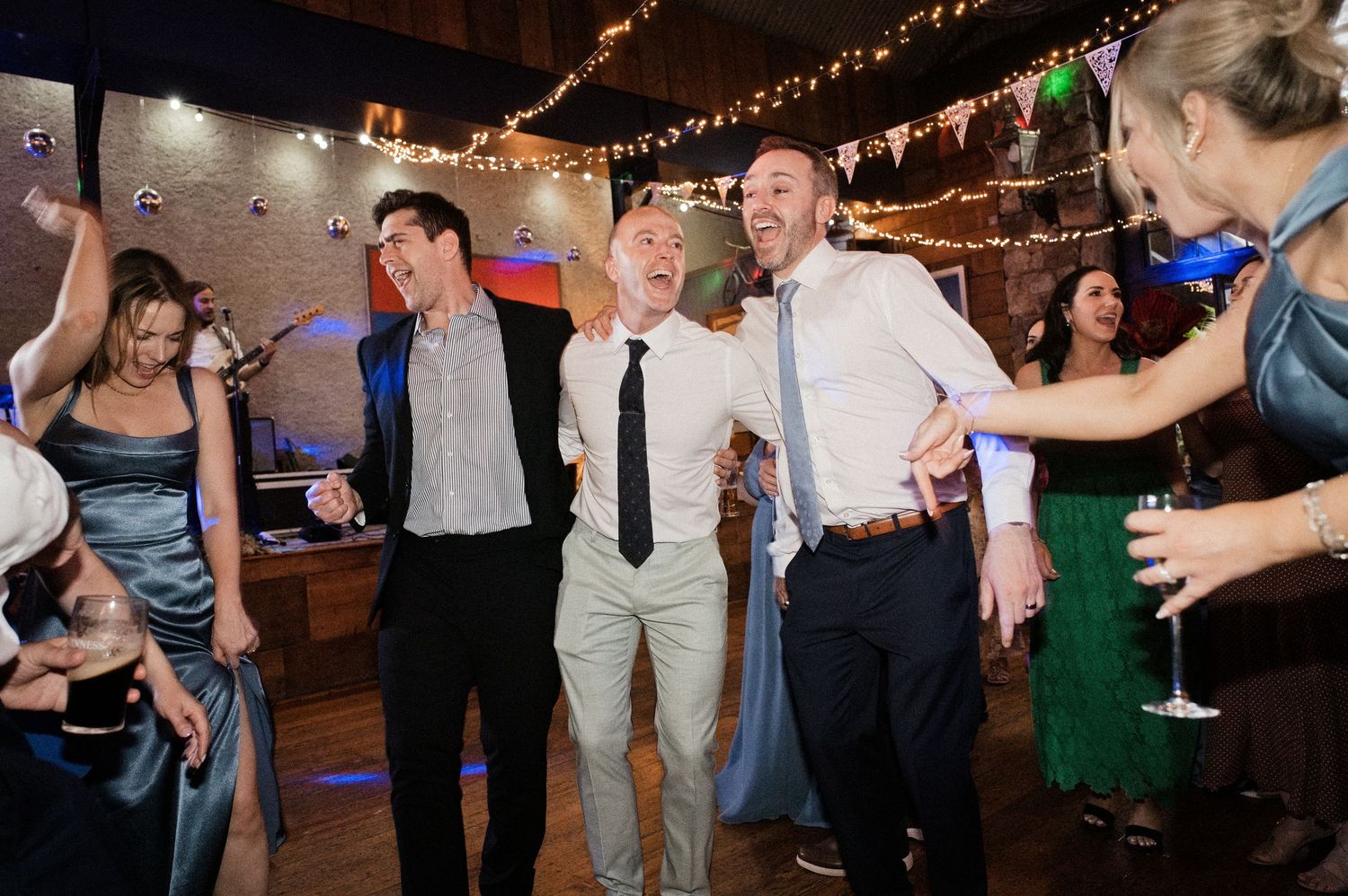 Guests dancing joyfully at a wedding reception in a warmly lit venue decorated with string lights.
