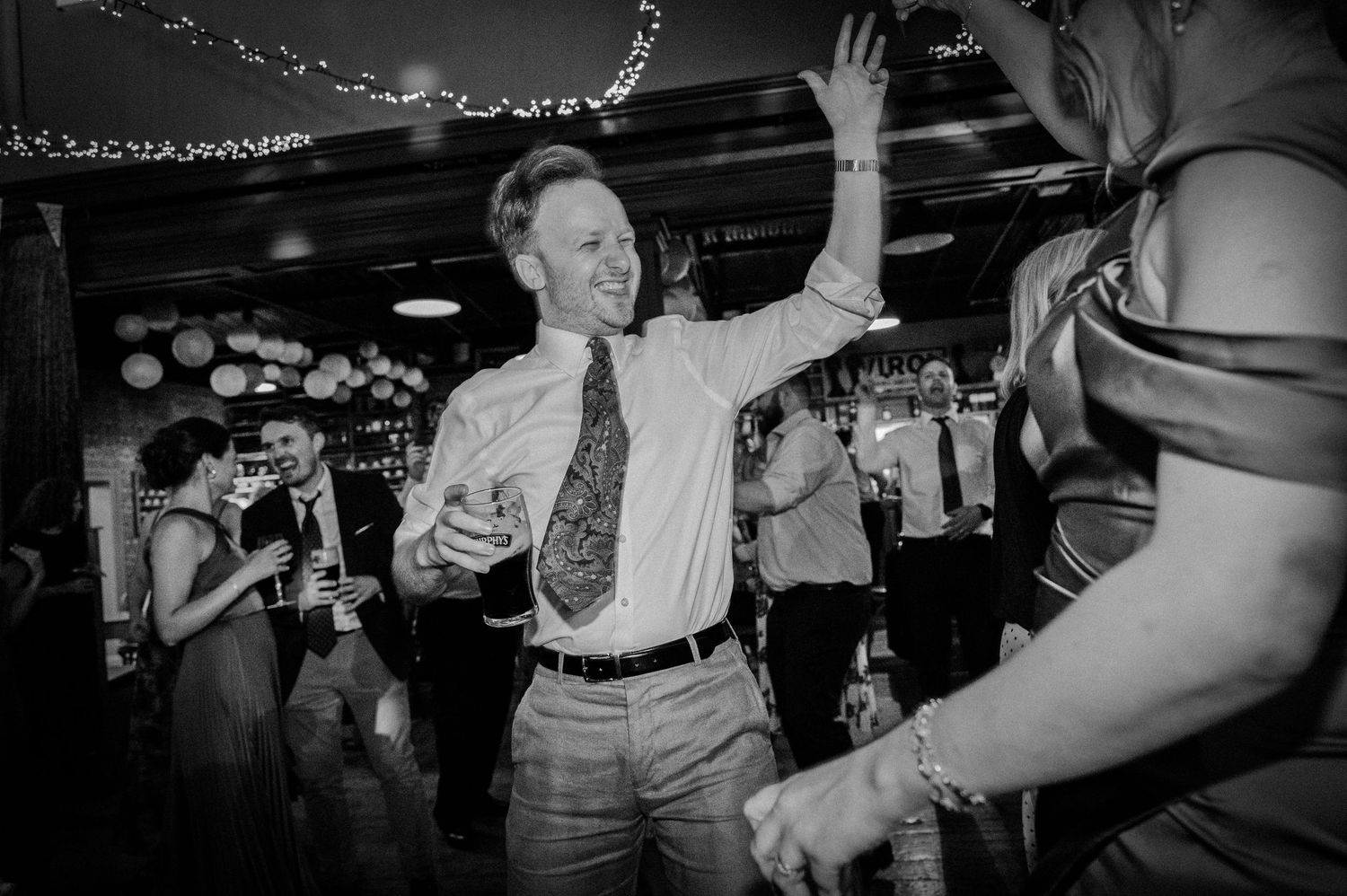 A joyful wedding guest dances and raises his hand while holding a drink at a lively wedding reception.