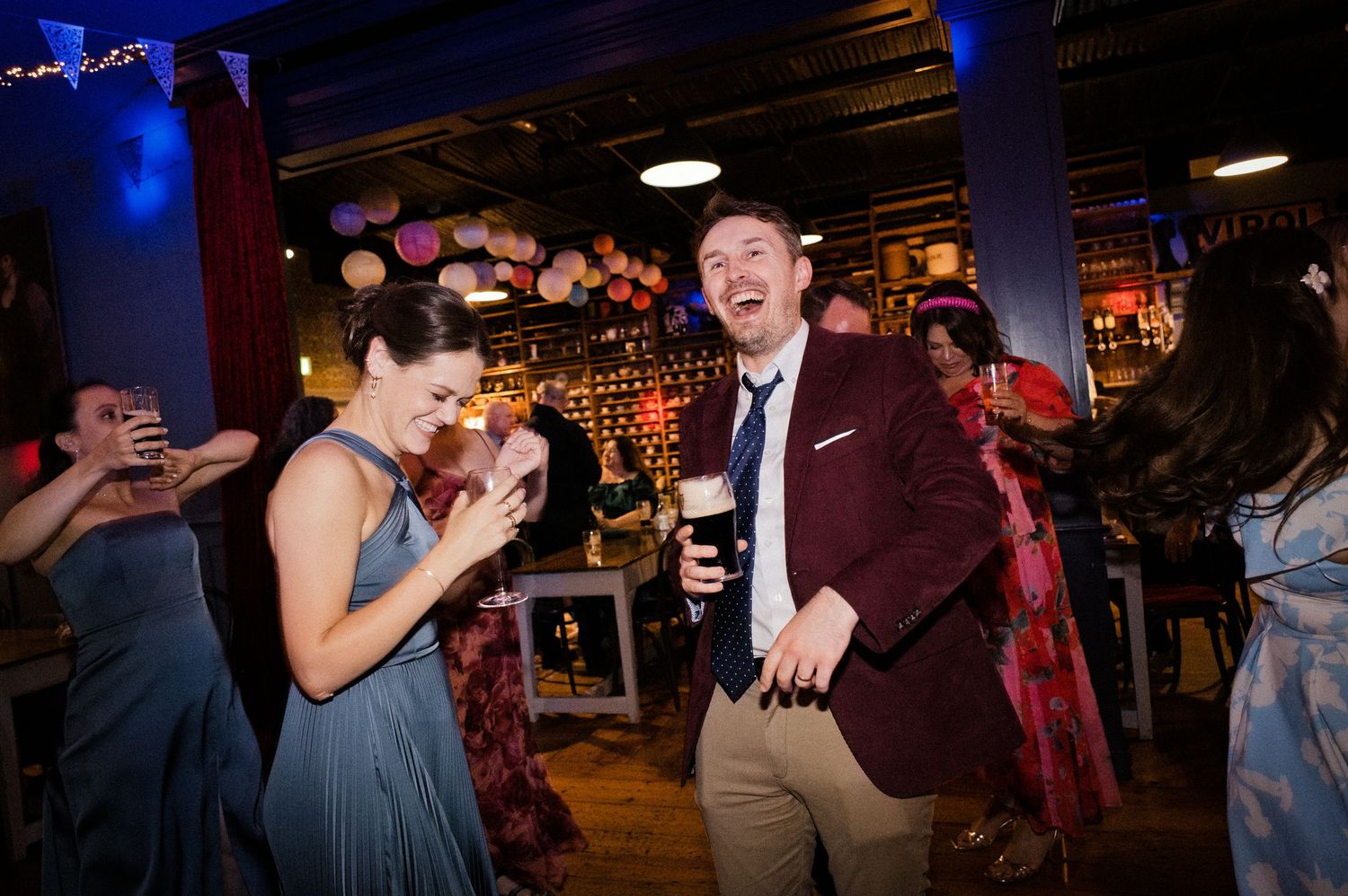 Couple laughing and dancing joyfully at a lively bar party, woman in blue dress, man in burgundy blazer holding a drink.