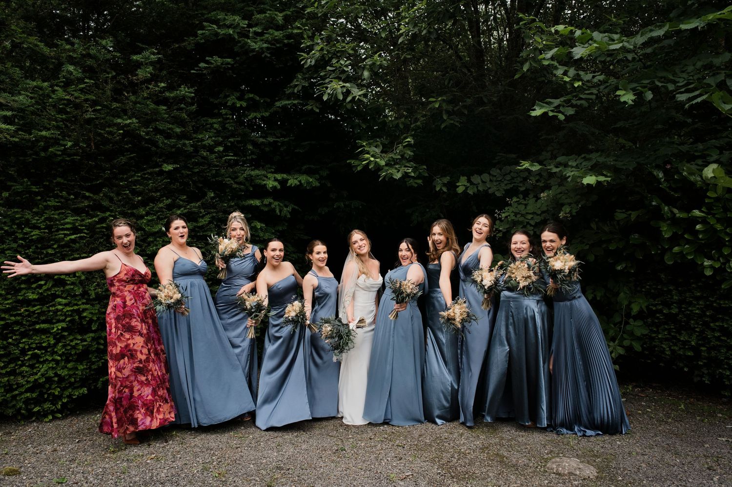 Bride poses with nine bridesmaids in dusty blue gowns holding bouquets outdoors.