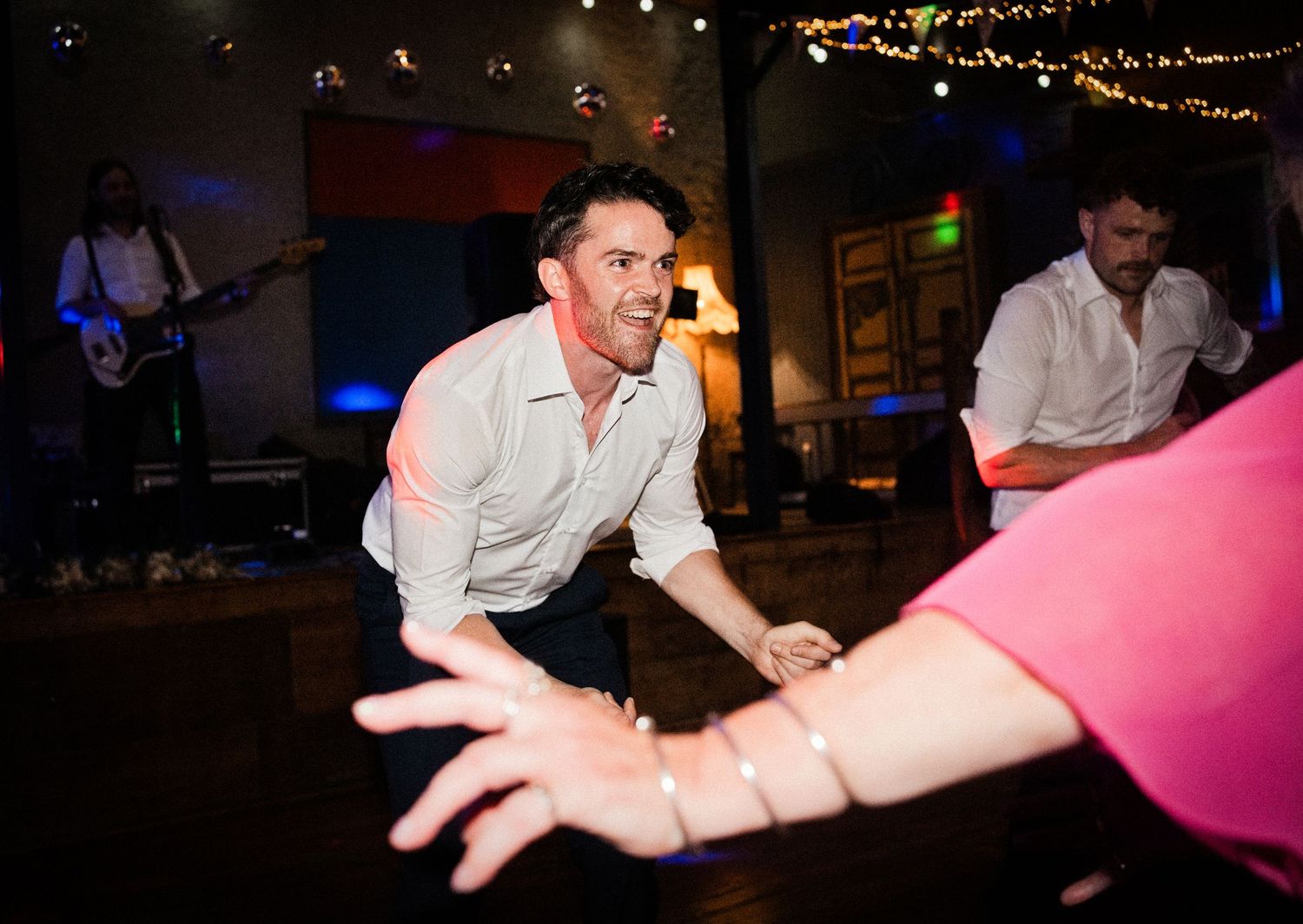A man enthusiastically reaches toward a guest on a lively wedding dance floor with a live band performing in the background.