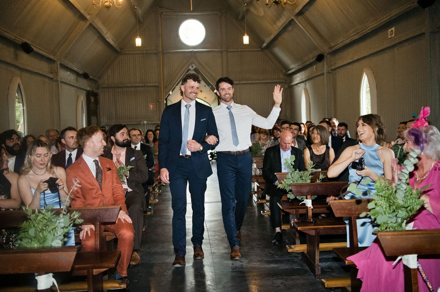 Two groomsmen walk joyfully down the aisle of a chapel as wedding guests celebrate around them.