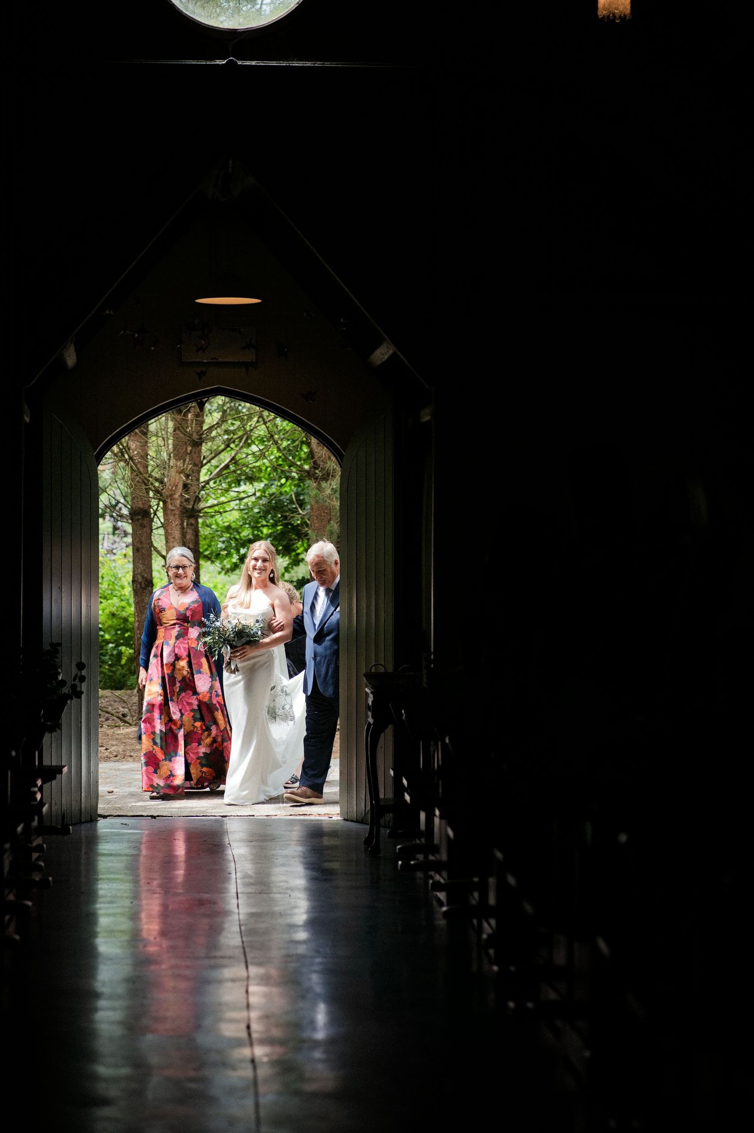 Bride flanked by family stands in sunlit church doorway before walking down the aisle.