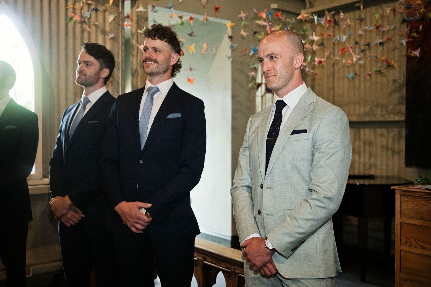 Three groomsmen in suits standing at wedding ceremony with colorful confetti decorations.
