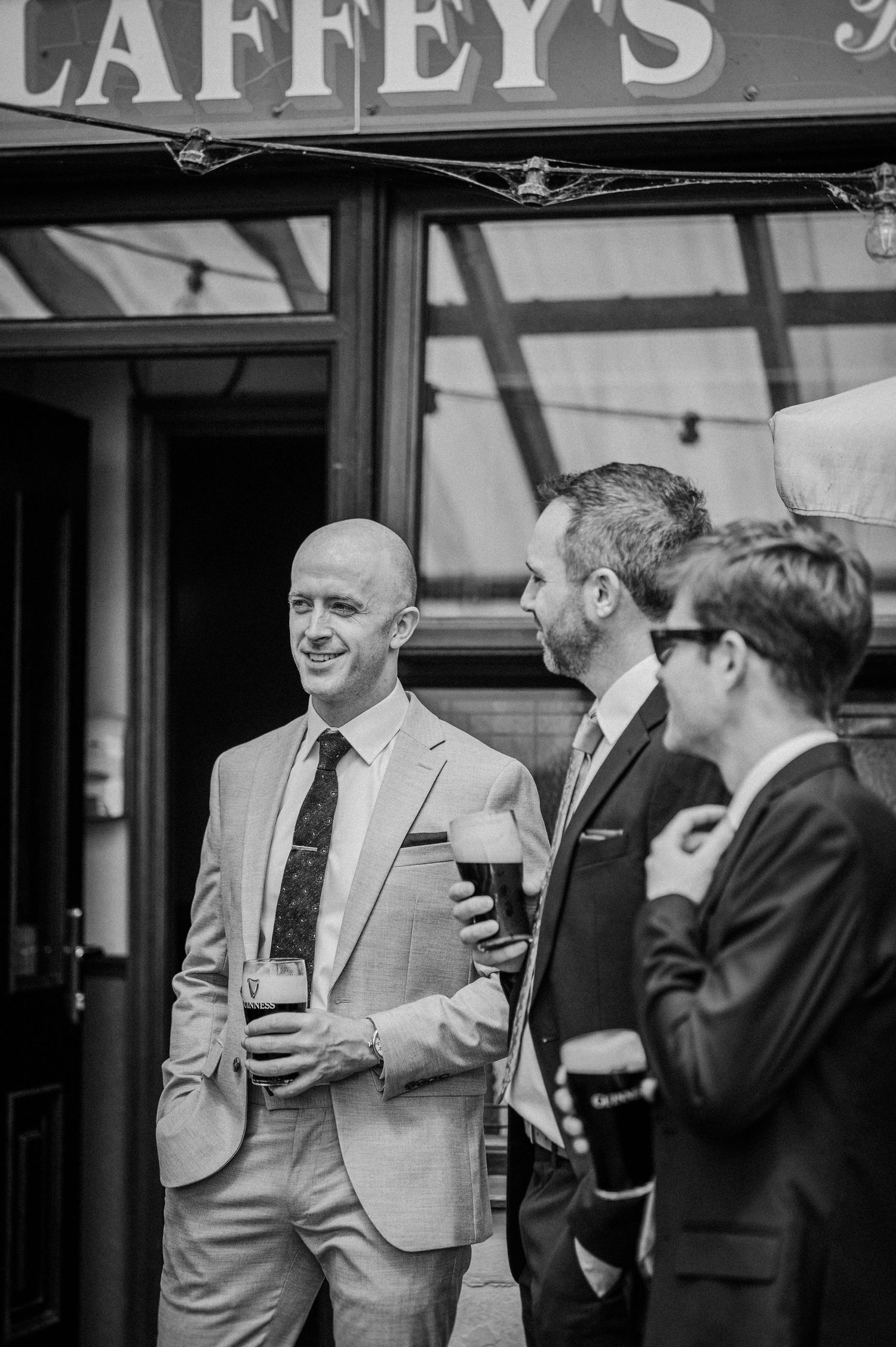 Three well-dressed men in suits enjoy Guinness pints outside Laffey's pub at a wedding celebration.