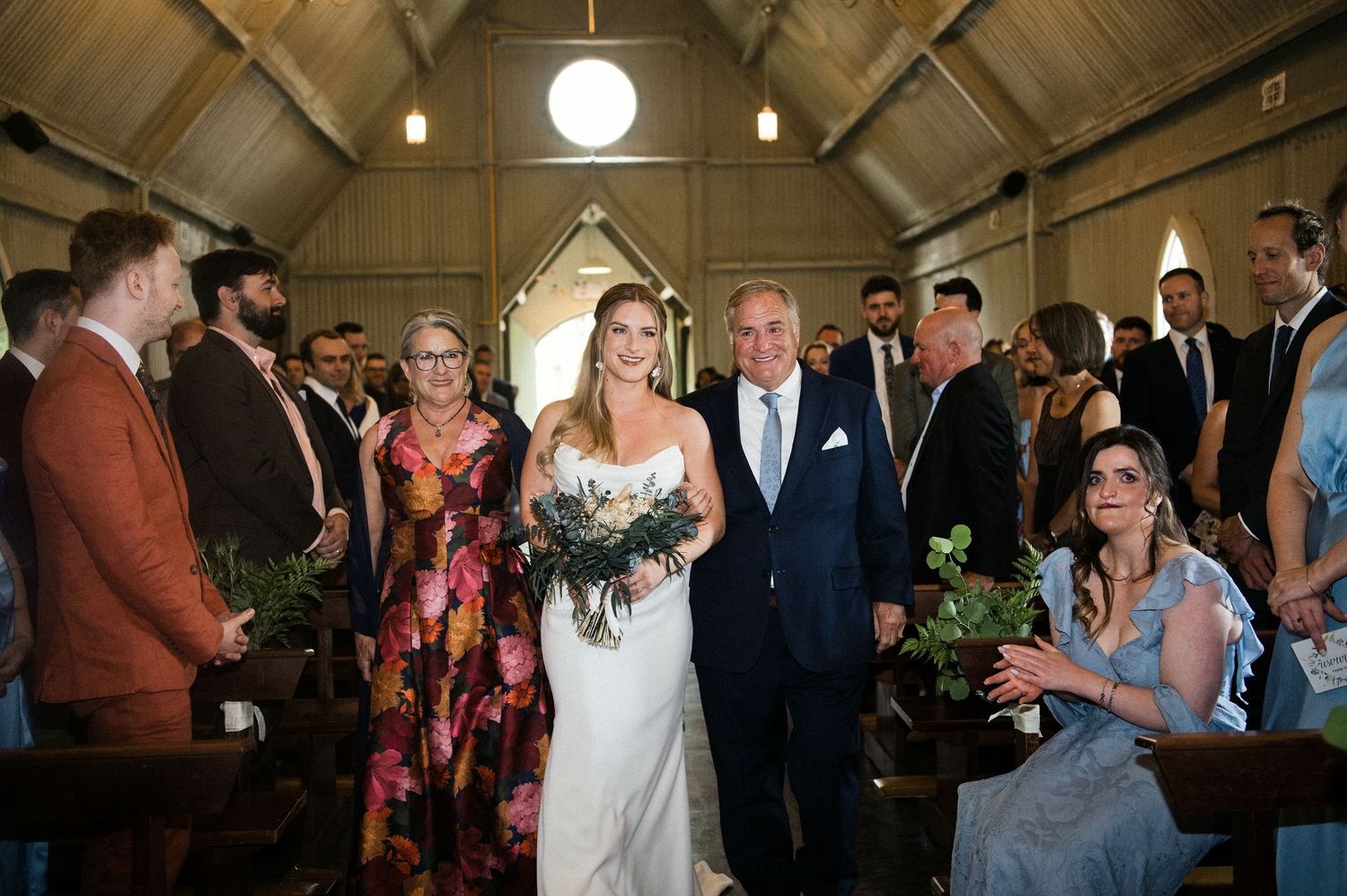 Bride walking down church aisle with father in floral-decorated rustic chapel surrounded by guests.