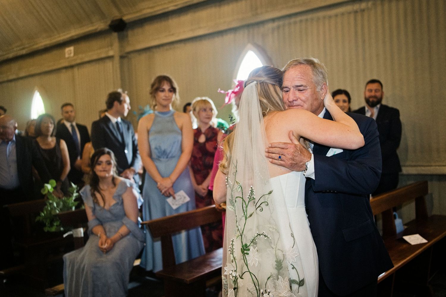 Emotional father-daughter hug at wedding ceremony altar with seated guests watching in church.