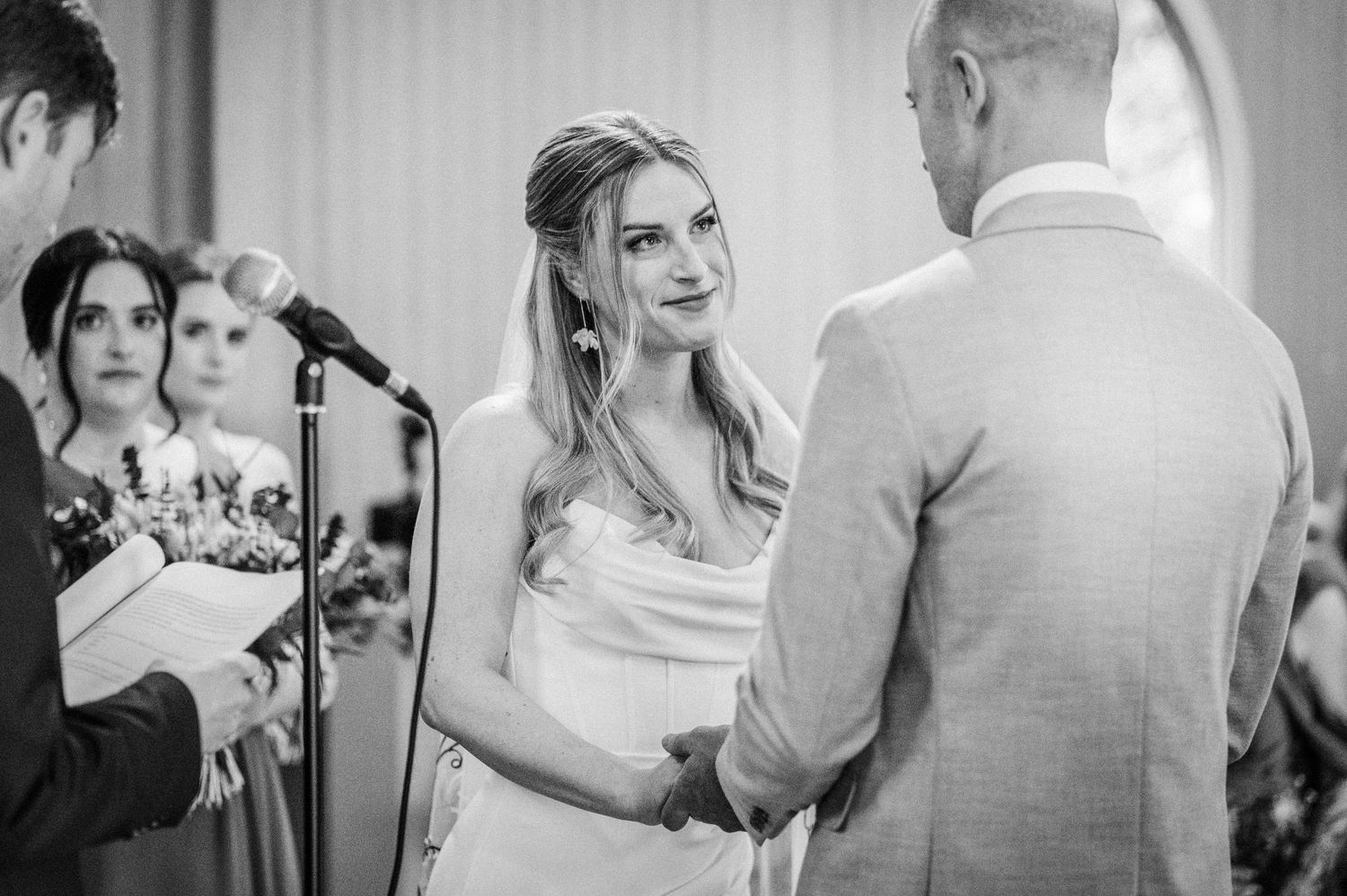 Bride smiles lovingly at groom during wedding ceremony vows at an elegant indoor venue.
