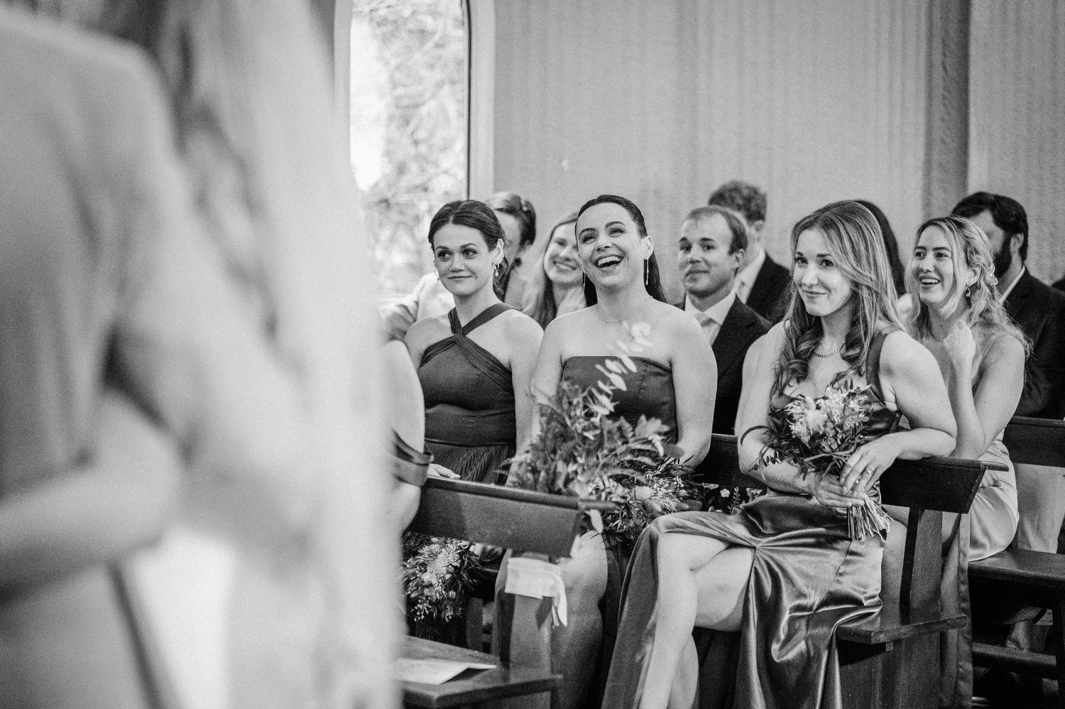 Smiling wedding guests seated in church pews during a ceremony, captured in black and white photography.