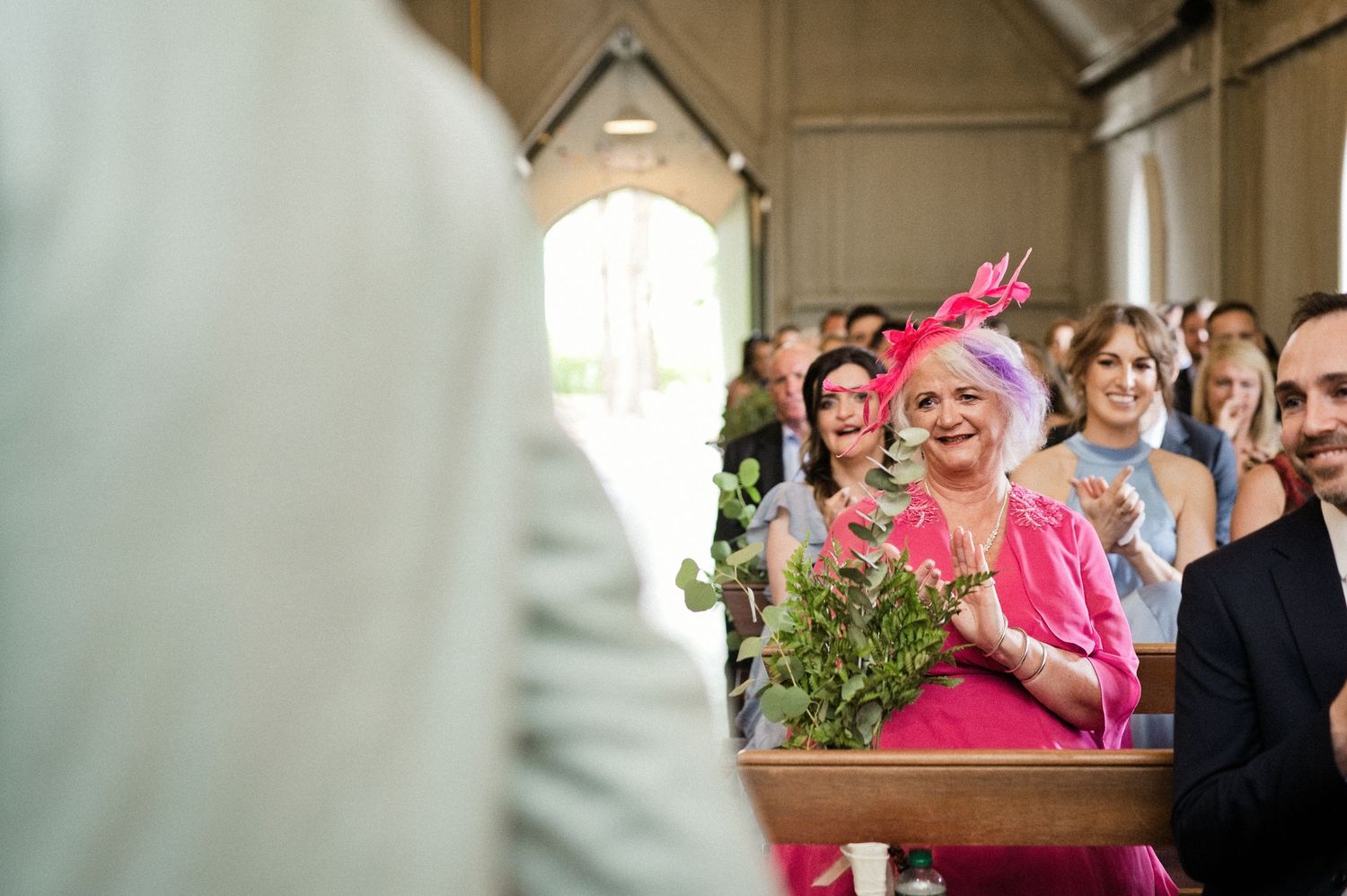 Emotional wedding guest in pink hat and dress smiling during a church ceremony while seated in wooden pews.