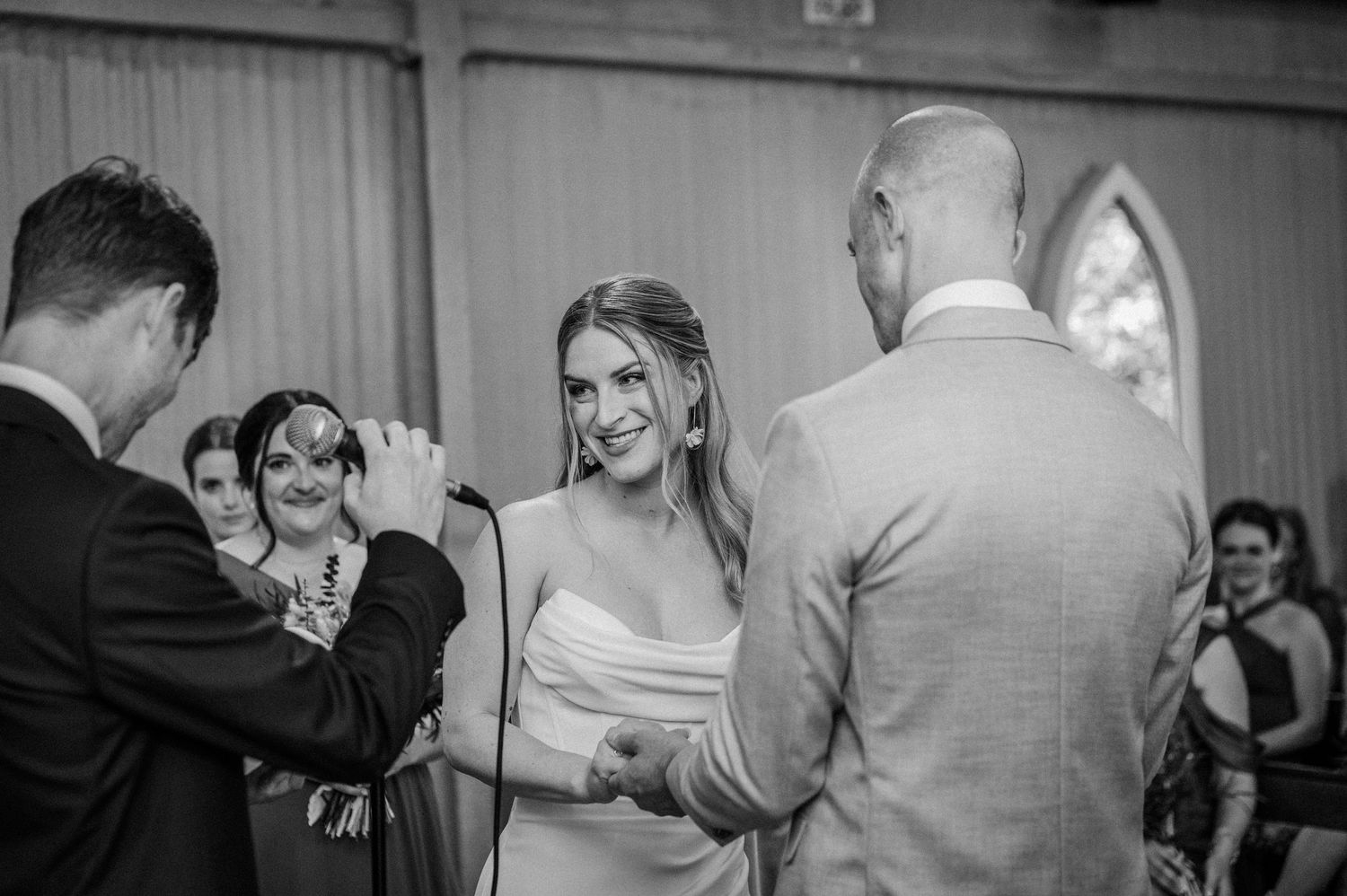 Bride smiling joyfully during wedding ceremony while exchanging vows with groom, black and white.