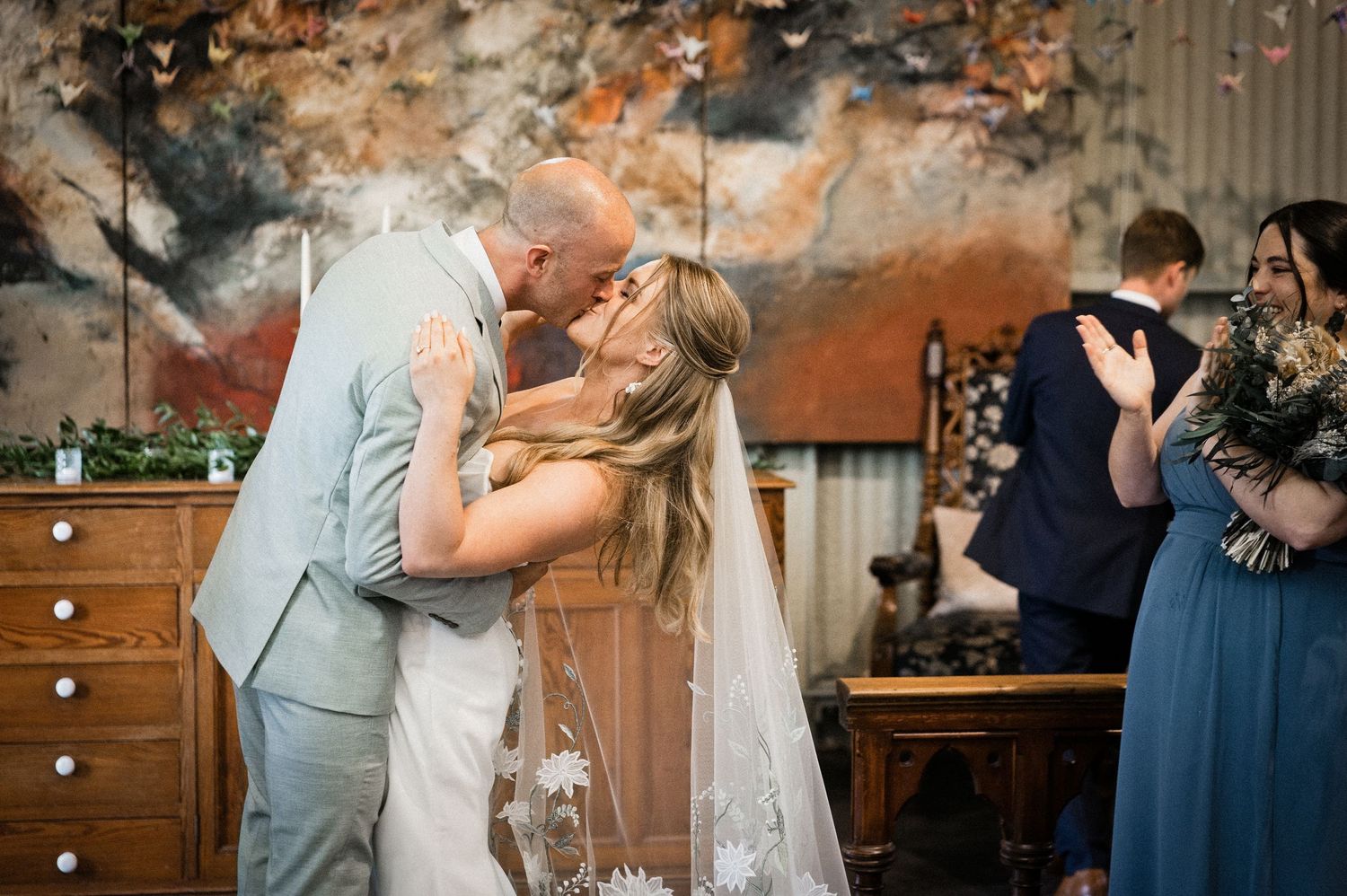 Newlyweds sharing first kiss at wedding altar with abstract painting backdrop and bridesmaid applauding.