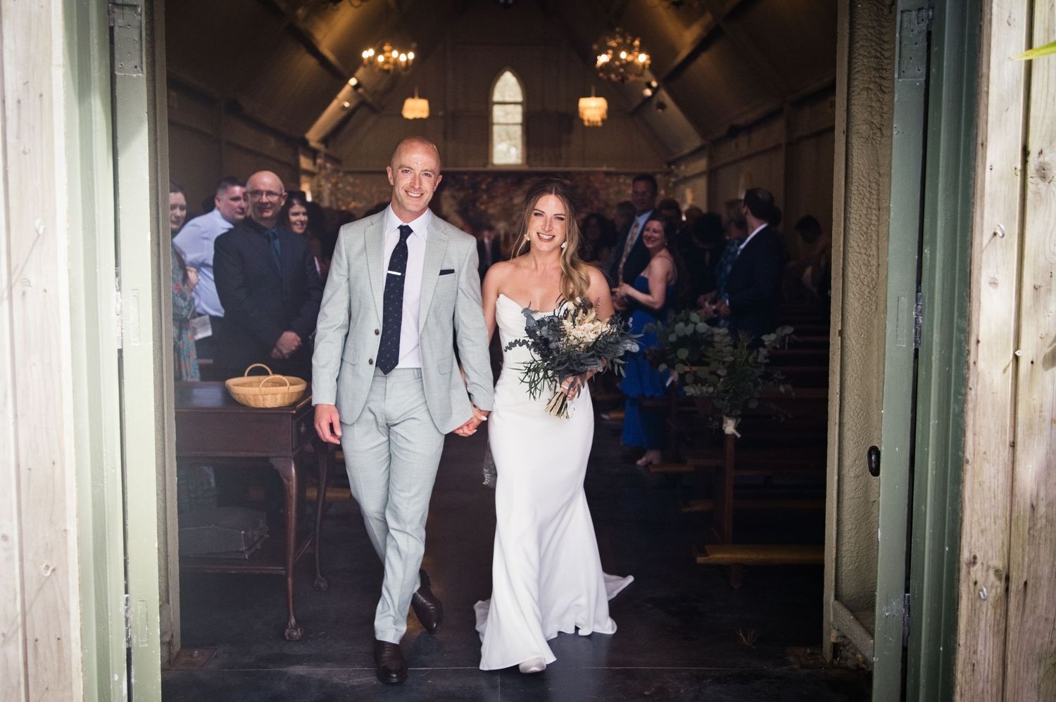 Newlyweds walking hand-in-hand back down aisle through church doors after wedding ceremony.