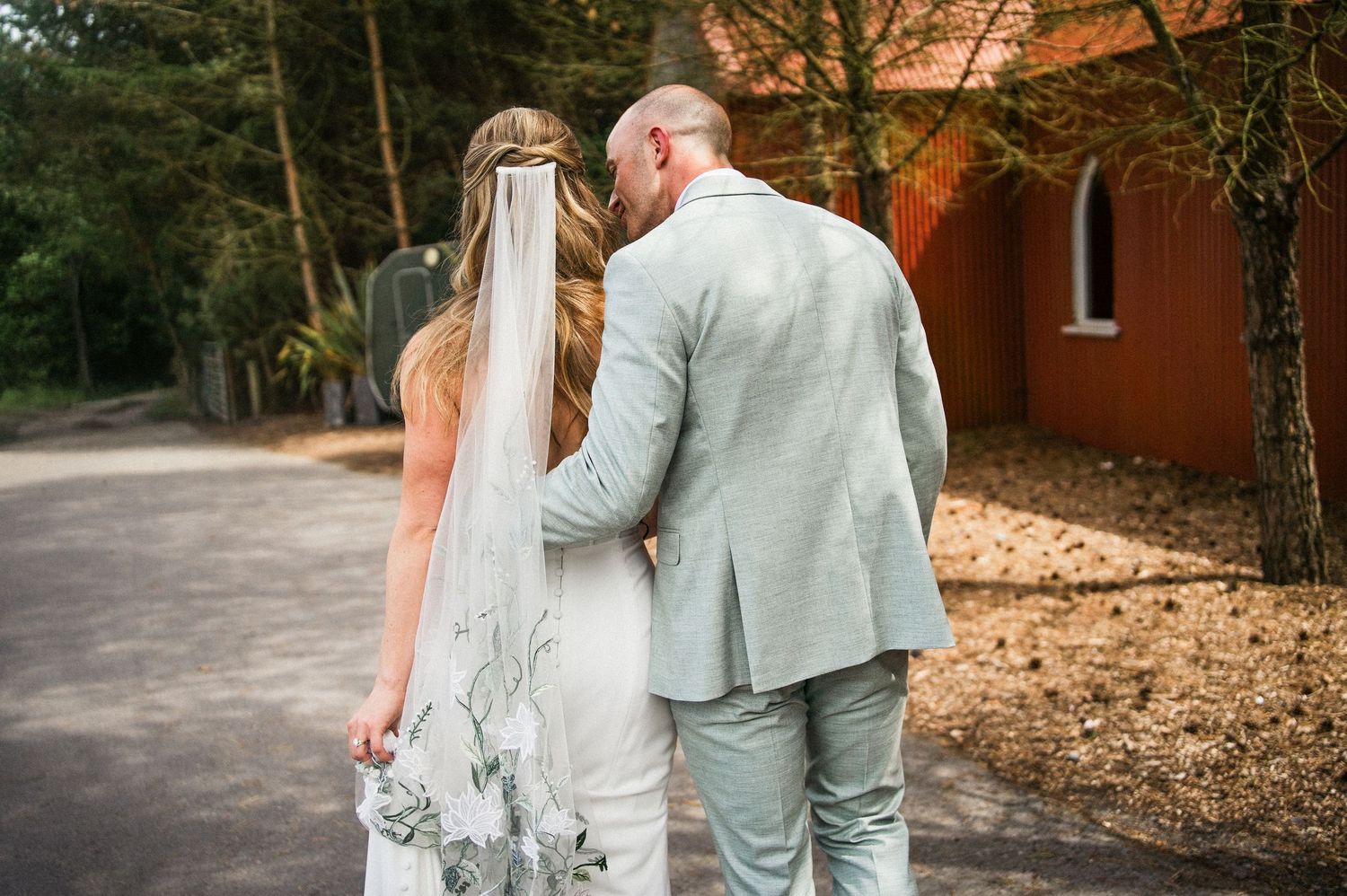 Bride and groom walking hand in hand from behind, bride wearing veil and groom in light grey suit.