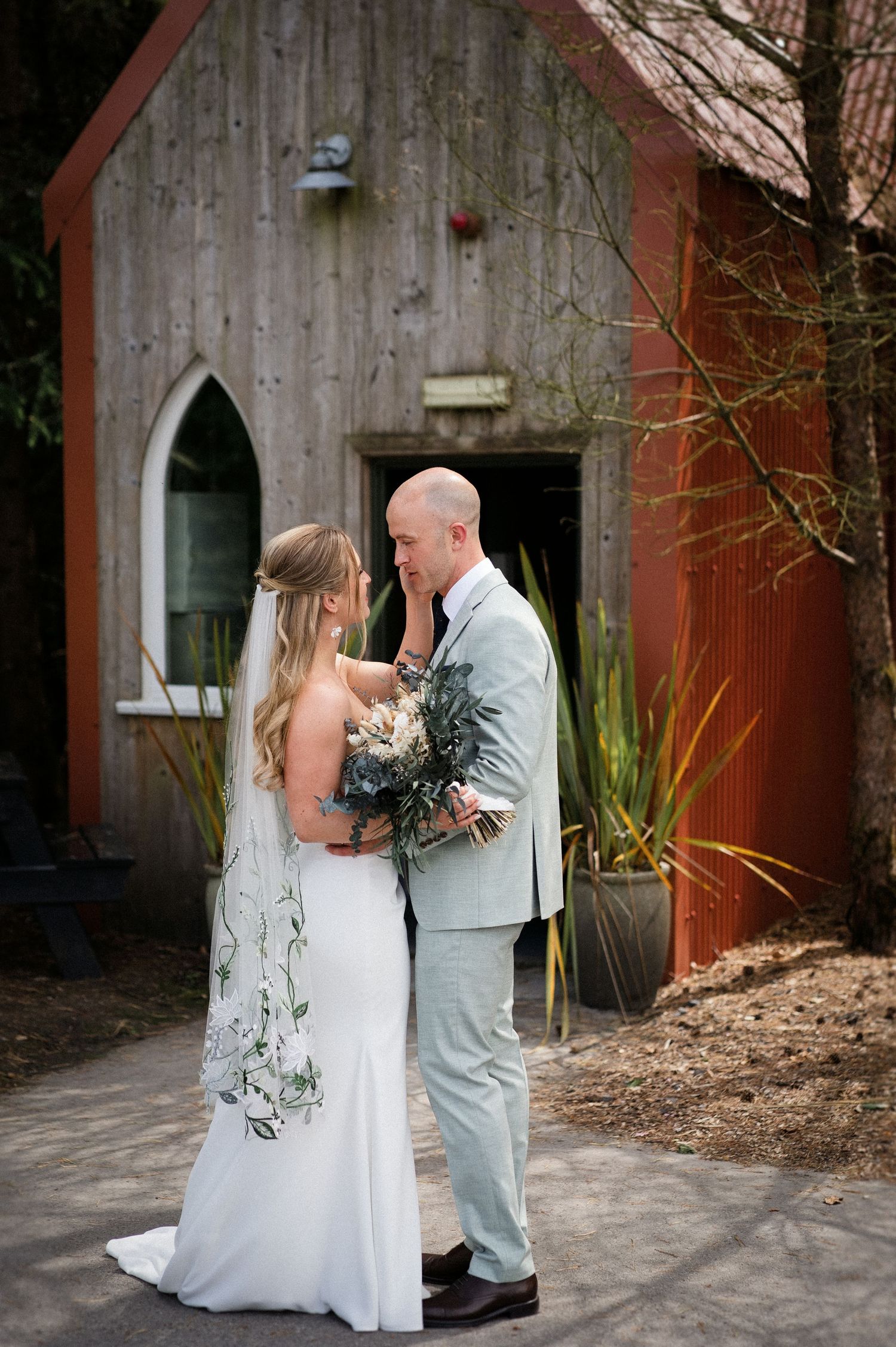 Bride and groom sharing an intimate moment outside a rustic wooden chapel, bride holding a greenery bouquet.